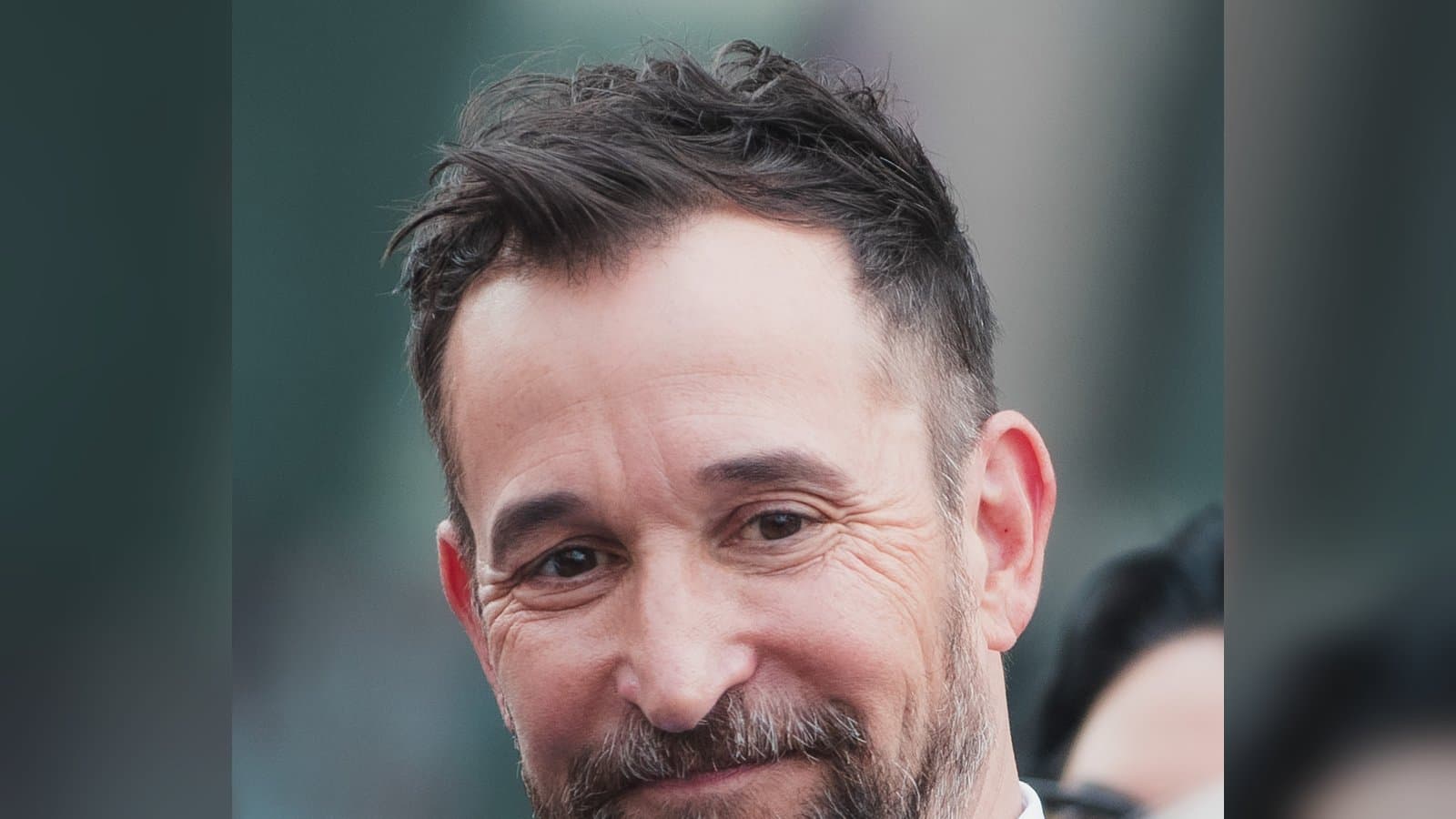 Noah Wyle, smiling with a salt-and-pepper beard, in a light blue jacket and open white shirt at his Hollywood Walk of Fame ceremony.