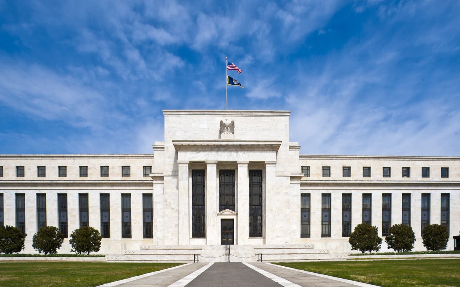 The white marble facade of the Federal Reserve's Eccles Building in Washington, with columns at its center entrance and a U.S. flag flying above it against a blue sky.
